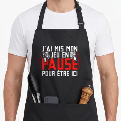 Tablier de cuisine noir avec inscription blanche et rouge J'ai mis mon jeu en pause pour être ici encadré par deux manettes de console.