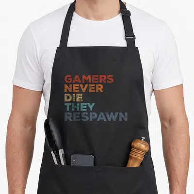 Tablier de cuisine noir avec inscription Gamers Never Die They Respawn en lettres multicolores dégradées.
