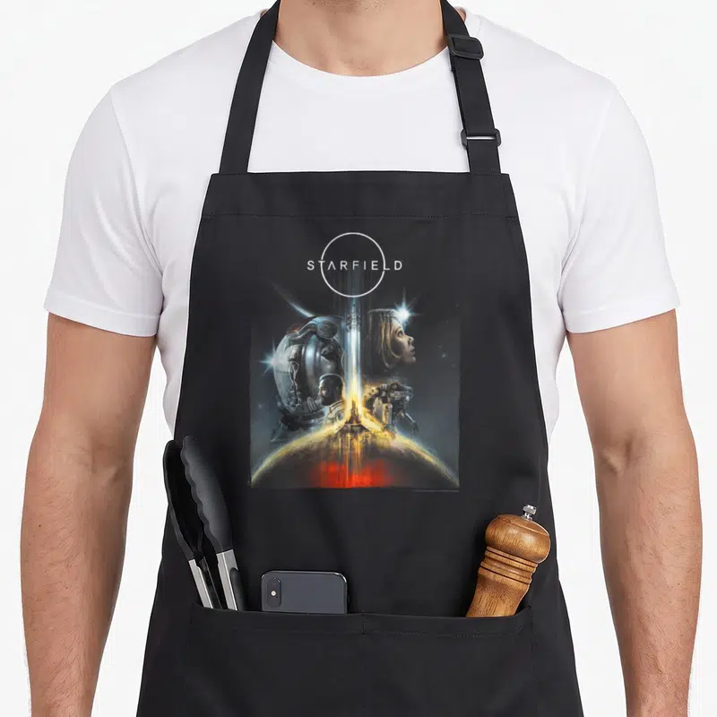 Tablier de cuisine noir avec illustration Starfield montrant des personnages et un vaisseau au-dessus d'une planète.