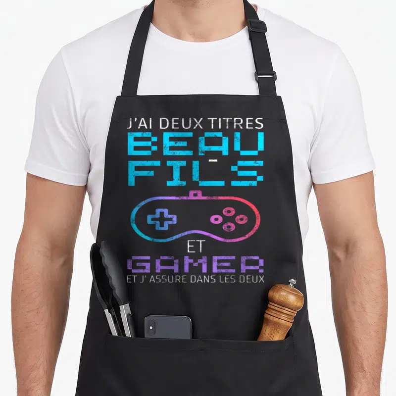 Tablier de cuisine noir avec texte J'ai deux titres beau-fils et gamer et une manette de jeu vidéo colorée au style pixelisé.