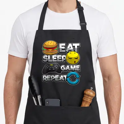 Tablier de cuisine noir avec illustration burger manette emoji sommeil et texte Mangez dormez jouez répétez en style cartoon coloré.
