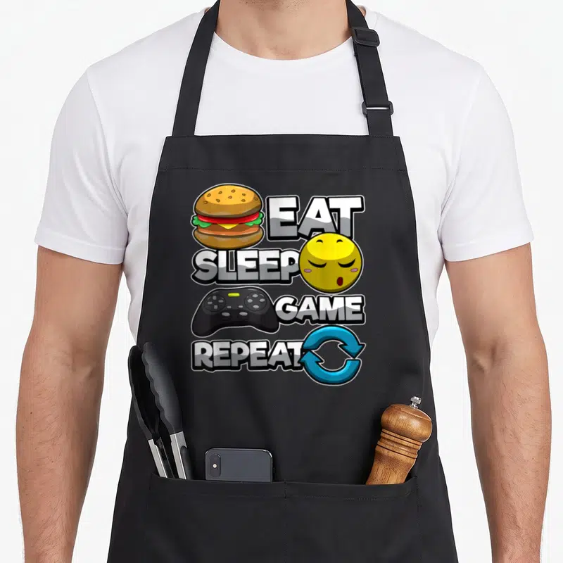 Tablier de cuisine noir avec illustration burger manette emoji sommeil et texte Mangez dormez jouez répétez en style cartoon coloré.