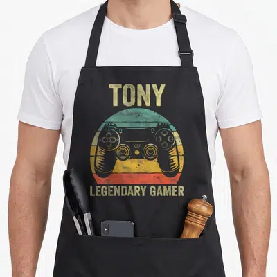 Tablier de cuisine noir avec une manette de jeu colorée et le texte Tony legendary gamer en jaune vieilli.