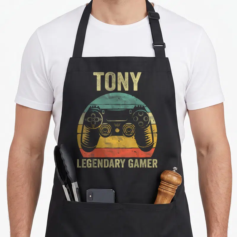 Tablier de cuisine noir avec une manette de jeu colorée et le texte Tony legendary gamer en jaune vieilli.