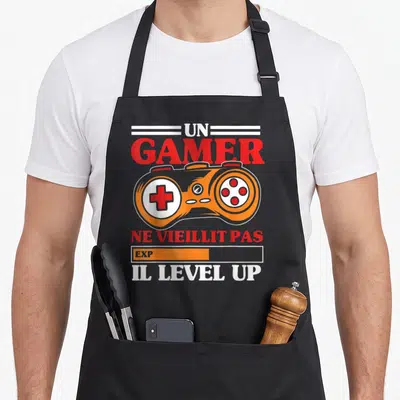 Tablier de cuisine noir avec manette orange et texte Un Gamer ne vieillit pas Il Level Up imprimé en rouge et blanc.