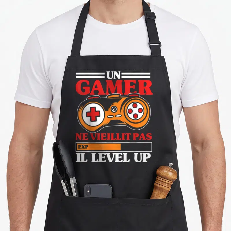 Tablier de cuisine noir avec manette orange et texte Un Gamer ne vieillit pas Il Level Up imprimé en rouge et blanc.