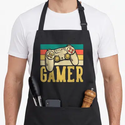 Tablier de cuisine noir avec illustration vintage d'une manette de jeu vidéo beige sur fond de bandes colorées et le mot GAMER en lettres jaunes.