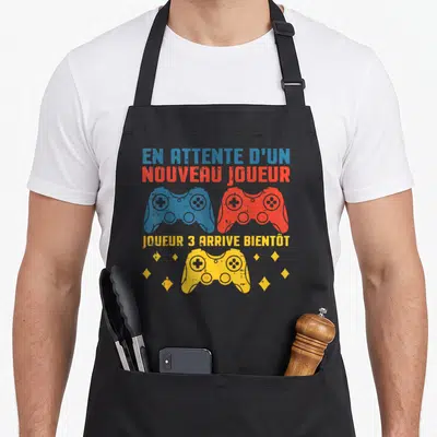 Tablier de cuisine noir avec trois manettes de jeu vidéo bleue, rouge et jaune et du texte humoristique.