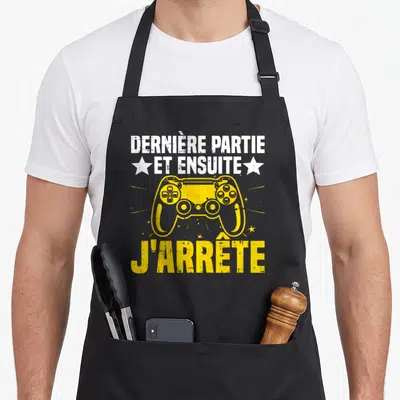 Tablier de cuisine noir avec inscription blanche et illustration de manette de console jaune au centre.
