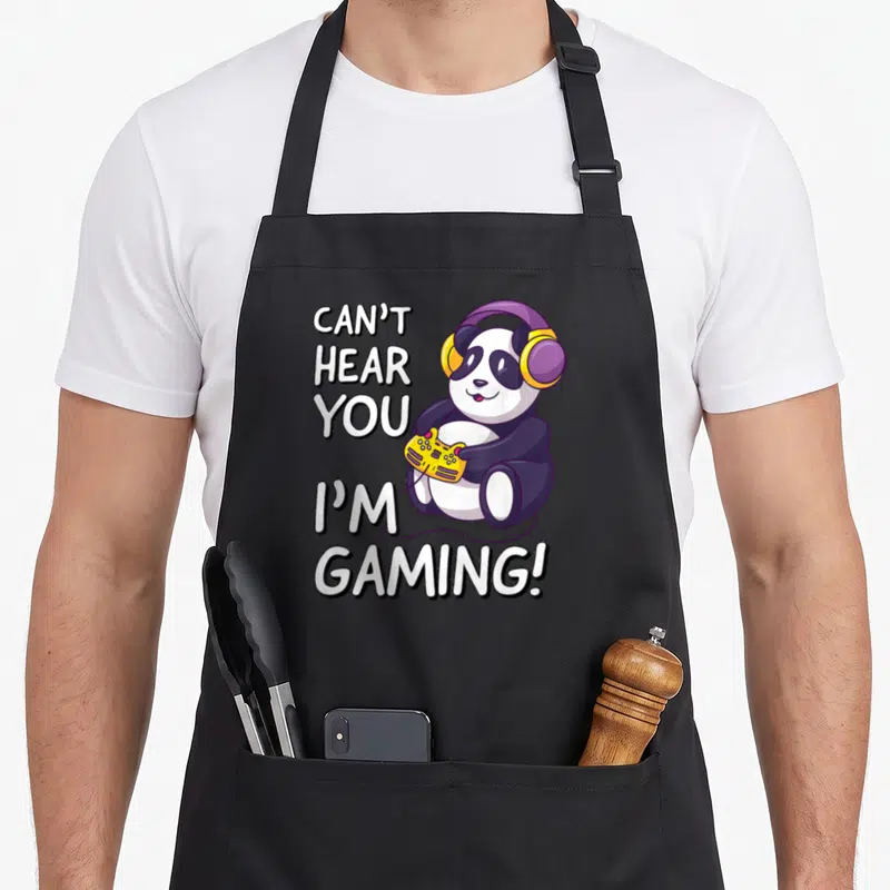 Tablier de cuisine noir avec un panda kawaii portant des écouteurs violet et tenant une manette jaune sous le texte Can't Hear You I'm Gaming.
