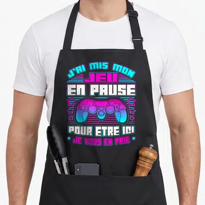 Tablier de cuisine noir avec une manette de jeu vidéo rose et bleue et du texte humoristique.