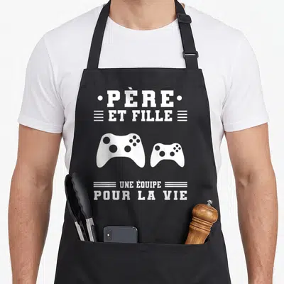 Tablier de cuisine noir avec texte blanc et deux manettes de jeu vidéo imprimés au centre.