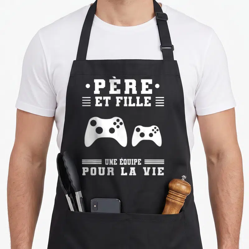 Tablier de cuisine noir avec texte blanc et deux manettes de jeu vidéo imprimés au centre.