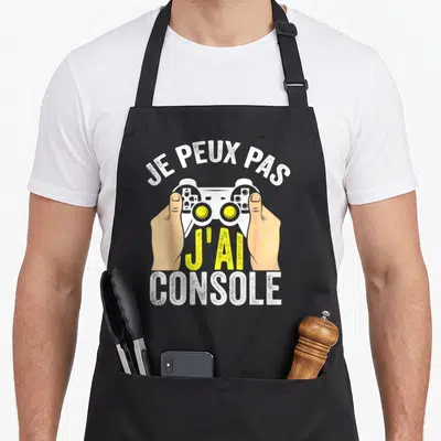 Tablier de cuisine noir avec illustration de mains tenant une manette blanche et texte Je peux pas j'ai console