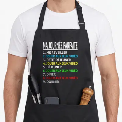 Tablier de cuisine noir avec liste colorée Ma Journée Parfaite Jouer aux Jeux Vidéo et Dormir imprimée au centre.