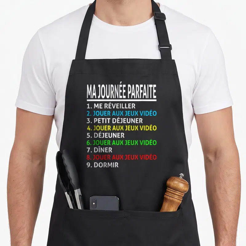 Tablier de cuisine noir avec liste colorée Ma Journée Parfaite Jouer aux Jeux Vidéo et Dormir imprimée au centre.