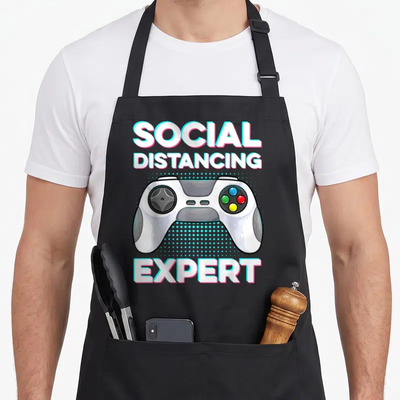 Tablier de cuisine noir avec une manette de jeu et le texte Social Distancing Expert en lettres colorées.