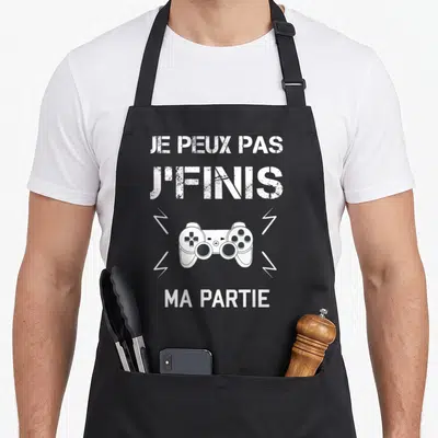 Tablier de cuisine noir avec une manette de jeu vidéo et le texte Je peux pas j'finis ma partie en blanc.