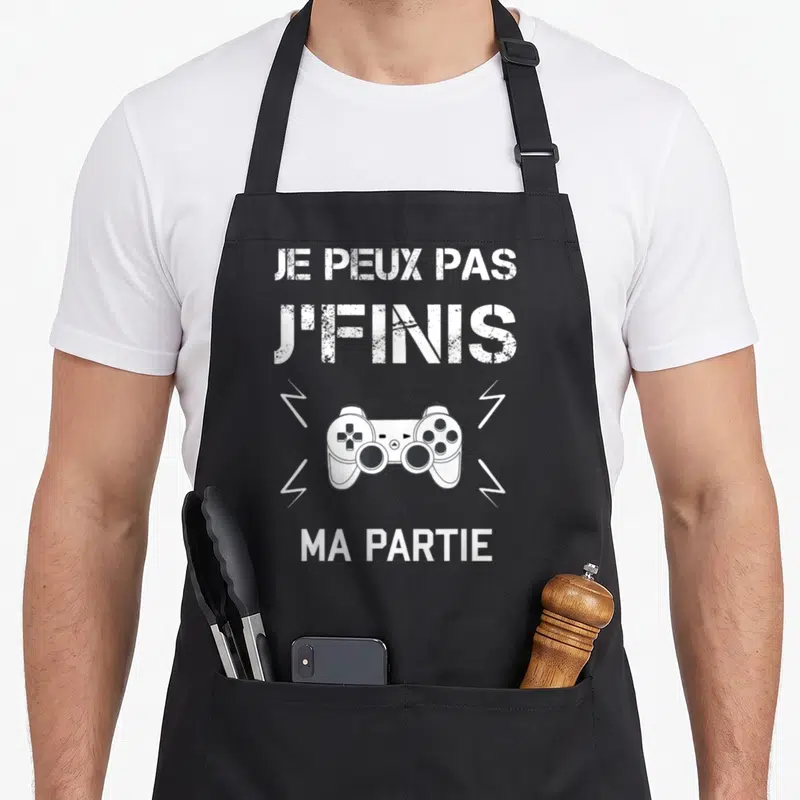 Tablier de cuisine noir avec une manette de jeu vidéo et le texte Je peux pas j'finis ma partie en blanc.