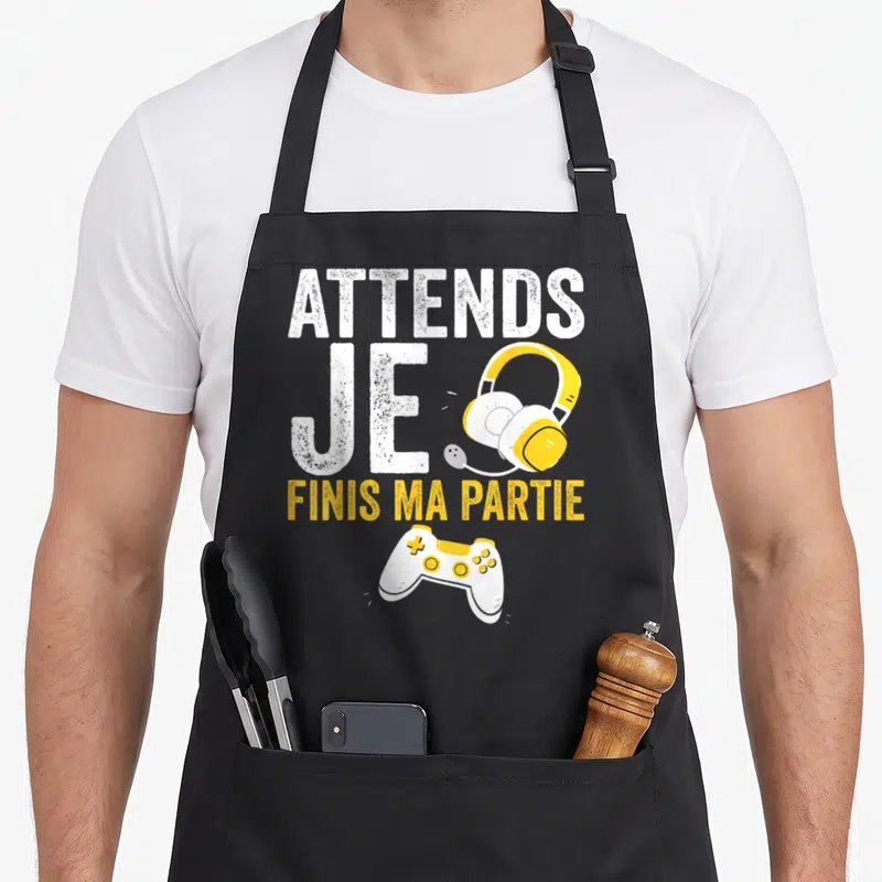 Tablier de cuisine noir avec inscription Attends Je Finis Ma Partie, casque jaune et manette de jeu blanche.