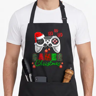 Tablier de cuisine noir avec une manette de jeu portant un bonnet de Père Noël et le texte Ho Ho Ho Gamer Christmas.
