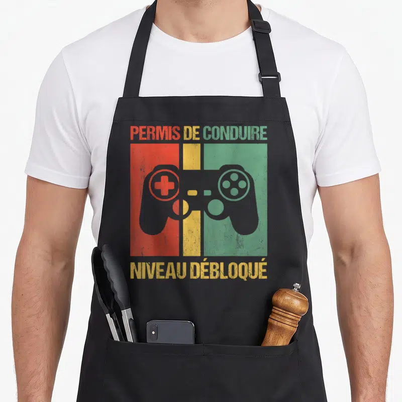 Tablier de cuisine noir avec illustration manette et texte Permis de conduire niveau débloqué sur fond tricolore vintage.