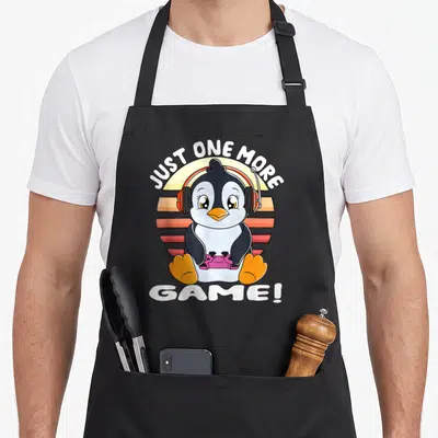Tablier de cuisine noir avec pingouin gamer portant un casque orange tenant une manette rose devant un soleil stylisé