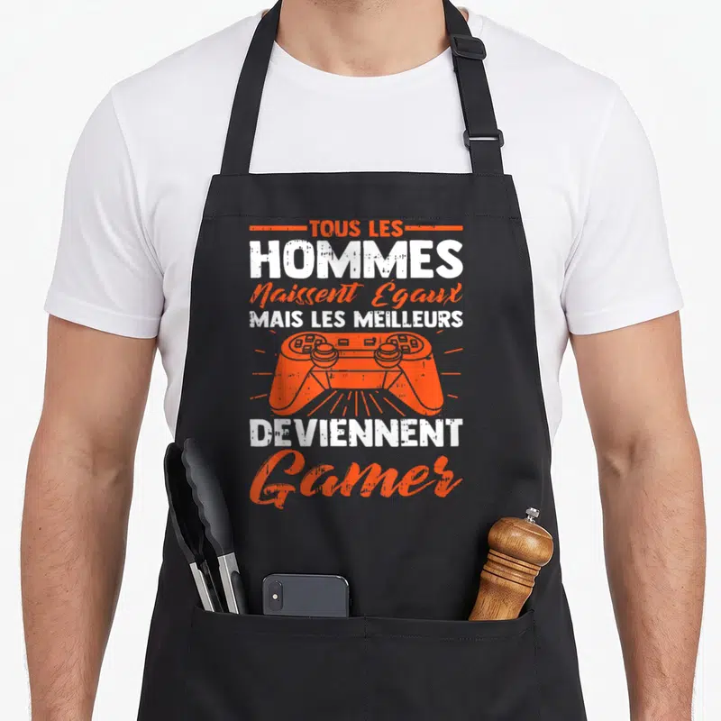 Tablier de cuisine noir avec texte blanc et orange et illustration de manette de jeu vidéo au centre du motif imprimé.