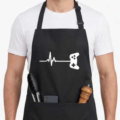 Tablier de cuisine noir avec une ligne de rythme cardiaque blanche se terminant par une silhouette de manette de jeu vidéo.