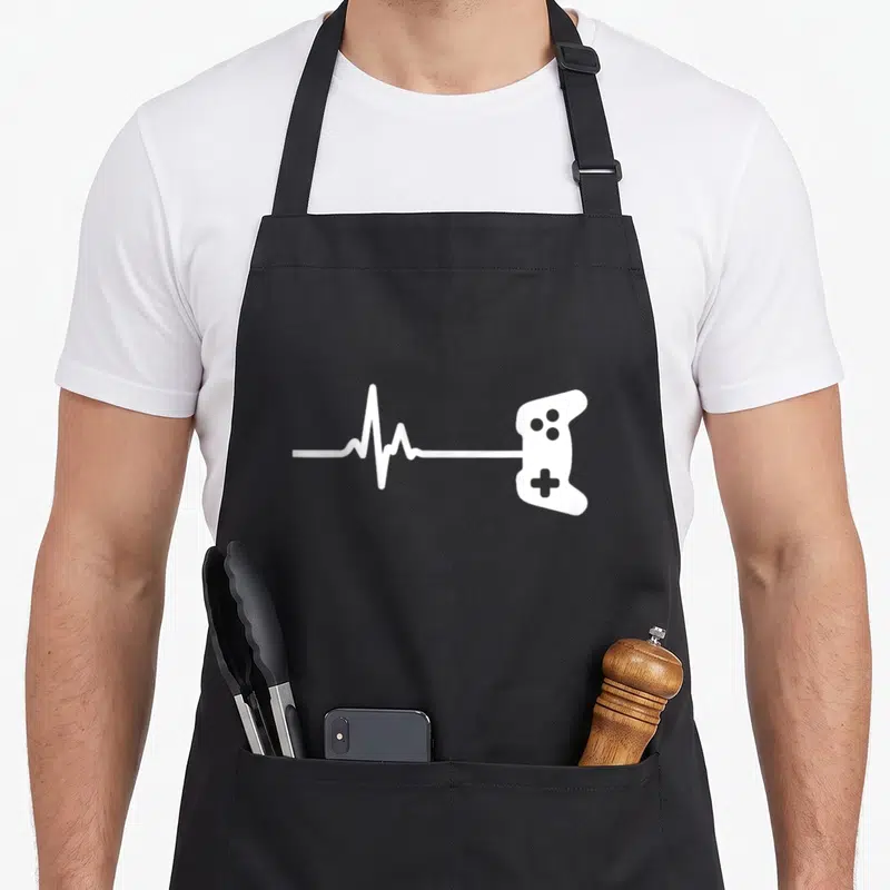 Tablier de cuisine noir avec une ligne de rythme cardiaque blanche se terminant par une silhouette de manette de jeu vidéo.