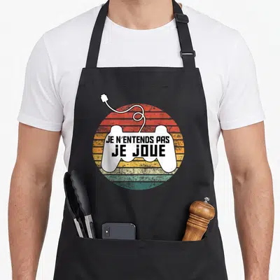 Tablier de cuisine noir avec une illustration de manette de jeu vidéo et le texte Je n'entends pas je joue sur fond coloré.