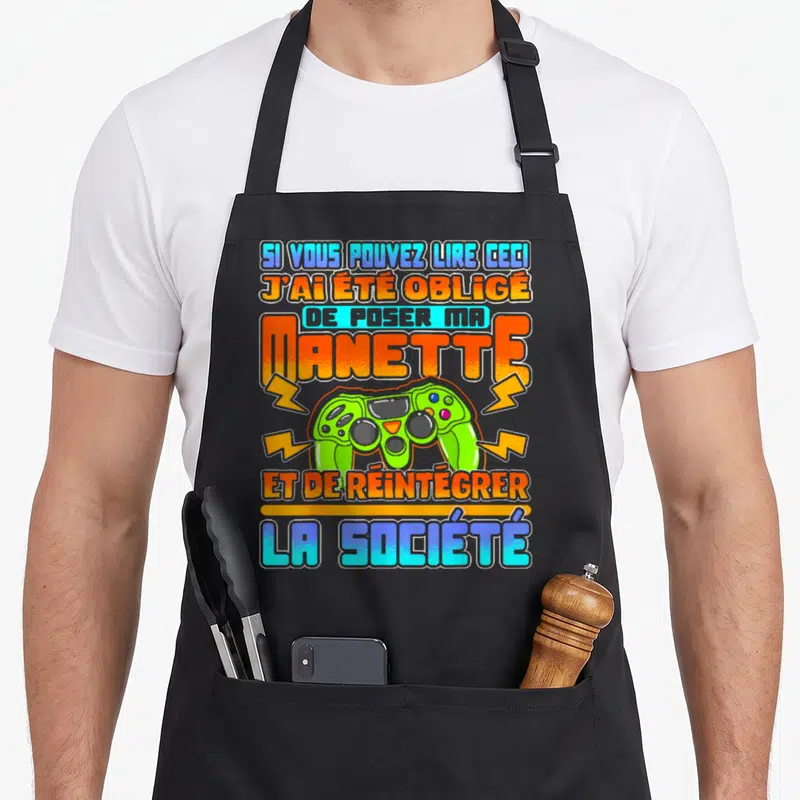Tablier de cuisine noir avec manette verte et texte humoristique sur le gaming et la société.
