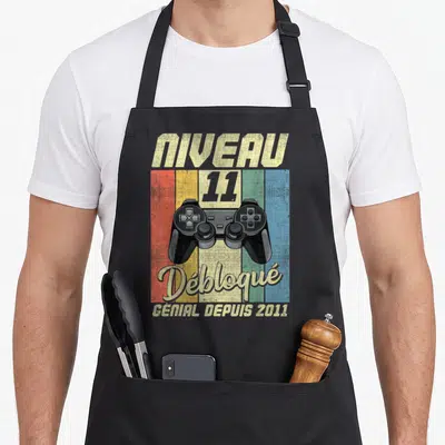 Tablier de cuisine noir avec illustration manette de jeu vidéo et texte Niveau 11 Débloqué Génial Depuis 2011 sur fond coloré.