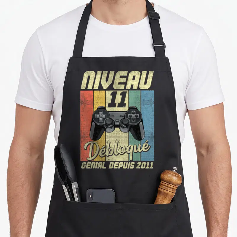 Tablier de cuisine noir avec illustration manette de jeu vidéo et texte Niveau 11 Débloqué Génial Depuis 2011 sur fond coloré.