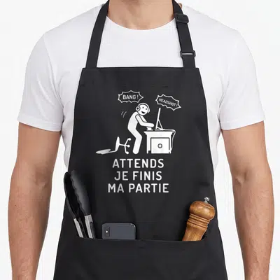 Tablier de cuisine noir avec illustration blanche d'un gamer au bureau et texte Attends je finis ma partie.