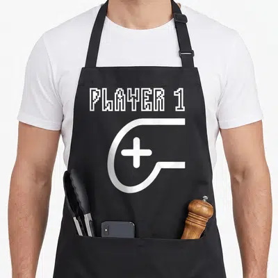 Tablier de cuisine noir avec texte Player 1 en pixel art et grande icône manette blanche stylisée.