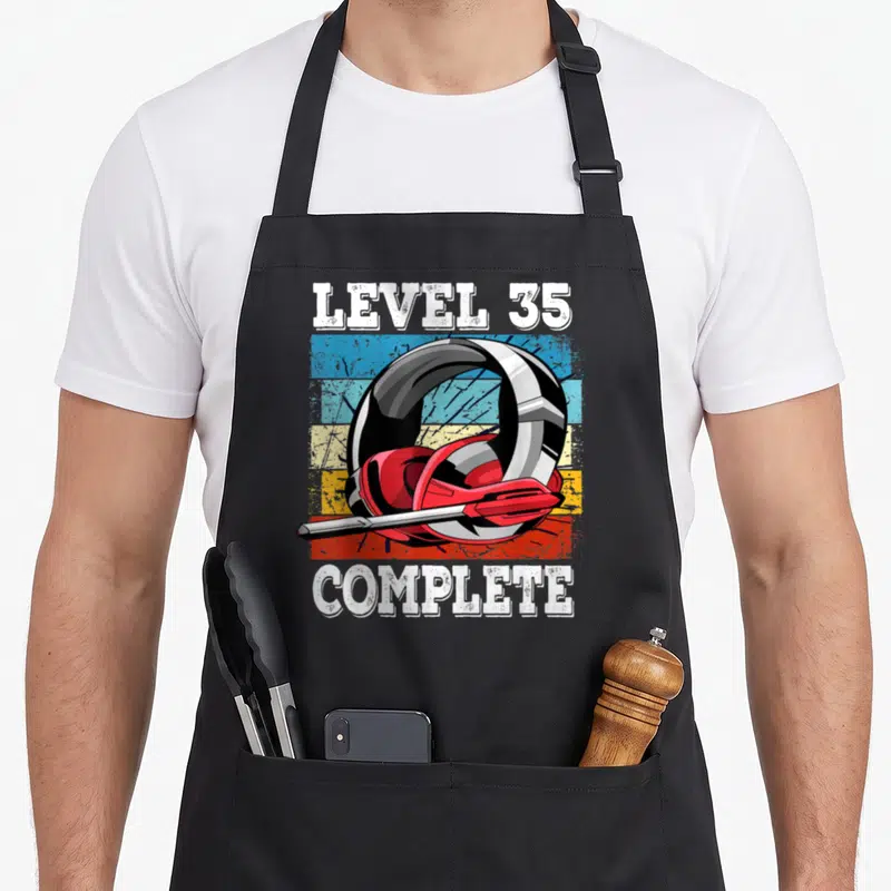 Tablier de cuisine noir avec illustration casque rouge et texte Level 35 Complete en style vintage.