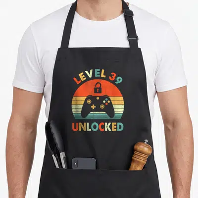 Tablier de cuisine noir avec illustration vintage manette de jeu et texte Level 39 Unlocked multicolore.