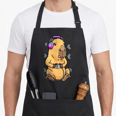 Tablier de cuisine noir avec illustration colorée d'un cabiaï gamer portant des écouteurs et tenant une manette blanche.