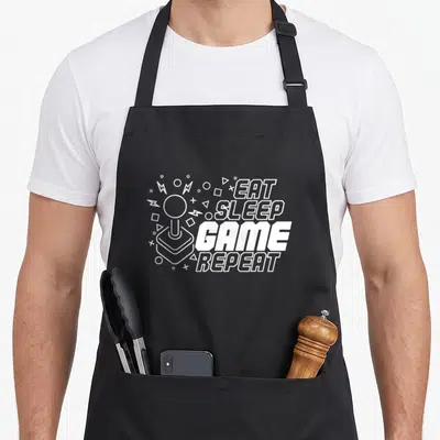 Tablier de cuisine noir avec inscription Eat Sleep Game Repeat et illustration de manette arcade blanche.