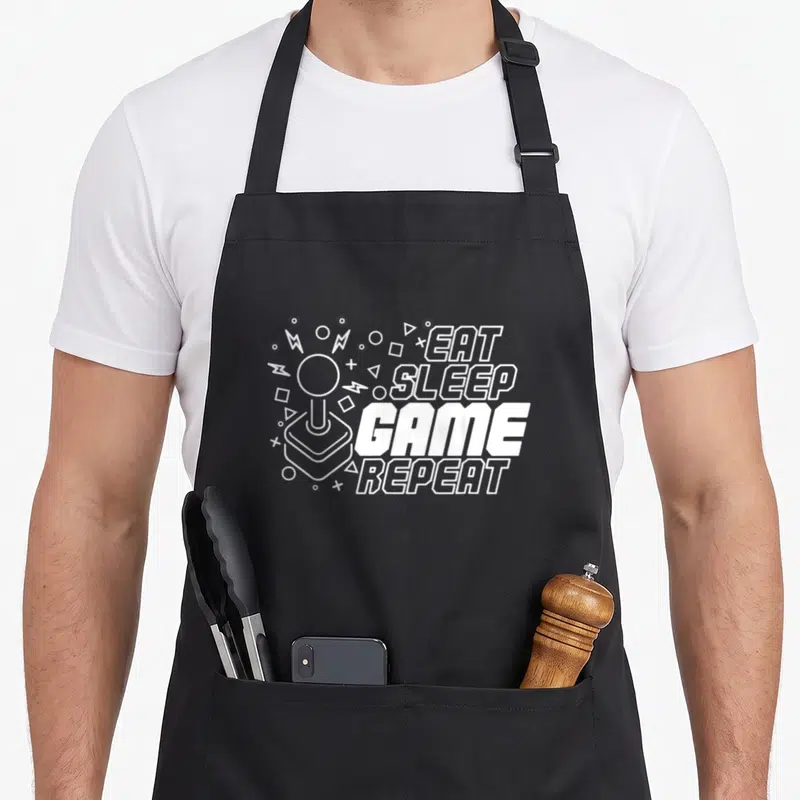 Tablier de cuisine noir avec inscription Eat Sleep Game Repeat et illustration de manette arcade blanche.