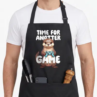Tablier de cuisine noir avec une loutre tenant une manette bleue et le texte Time for Anotter Game en blanc.
