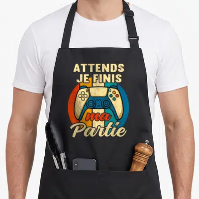 Tablier de cuisine noir avec illustration de manette de jeu et texte Attends Je Finis Ma Partie sur fond coloré.