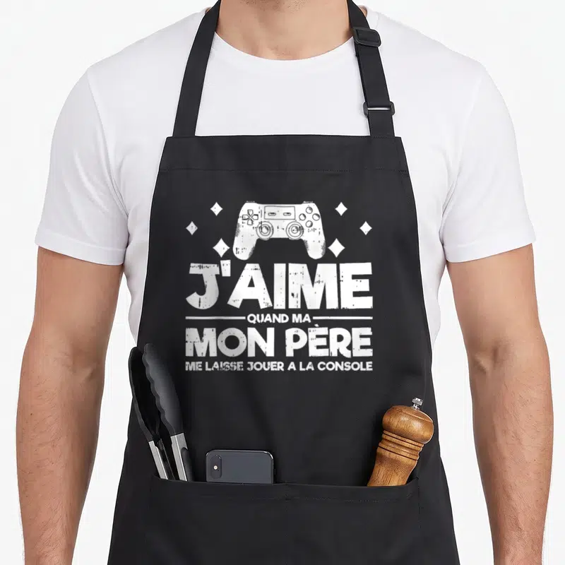 Tablier de cuisine noir avec une illustration de manette de jeu et le texte J'aime quand mon père me laisse jouer à la console.