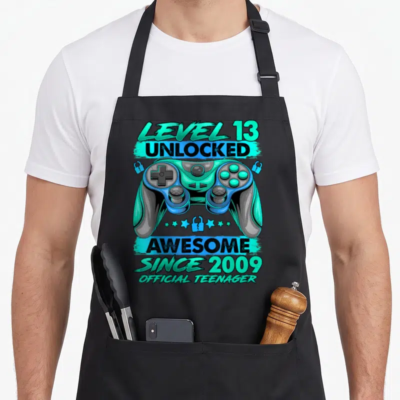 Tablier de cuisine noir avec illustration de manette bleue et texte Level 13 Débloqué Génial Depuis 2009 Official Teenager.