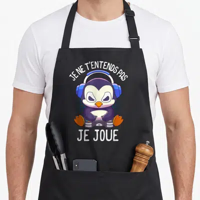 Tablier de cuisine noir avec manchot portant des écouteurs et tenant une manette, texte blanc au-dessus et en dessous.