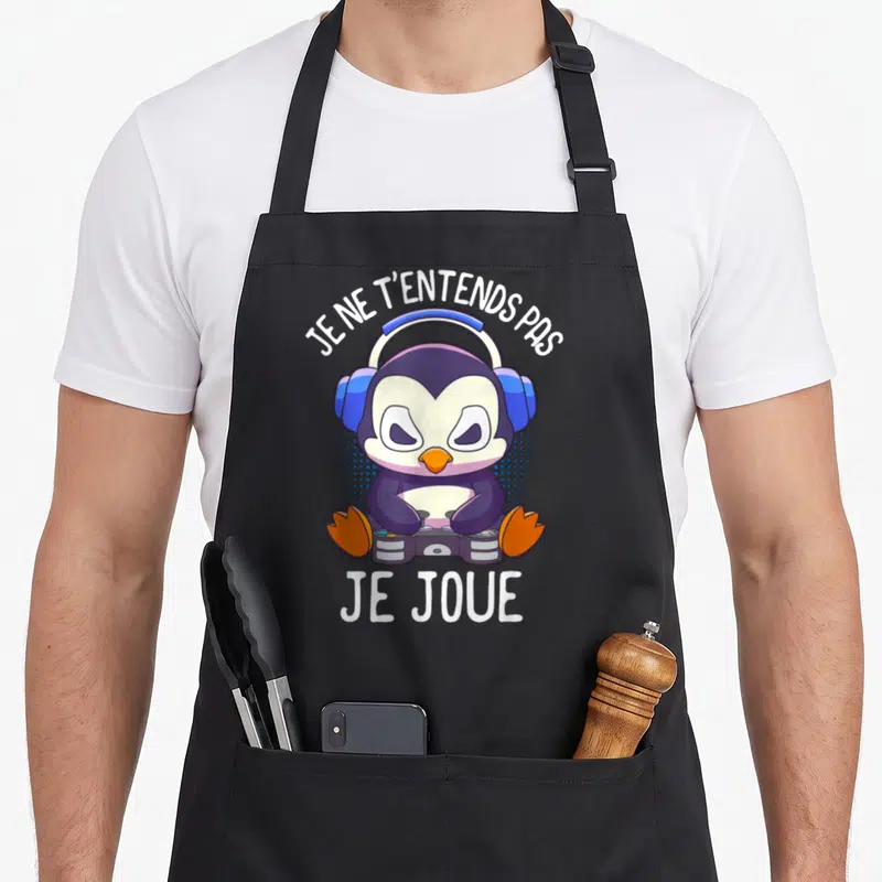 Tablier de cuisine noir avec manchot portant des écouteurs et tenant une manette, texte blanc au-dessus et en dessous.