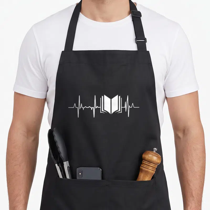 Tablier de cuisine noir avec un motif blanc montrant un livre ouvert au centre d'une ligne de rythme cardiaque.