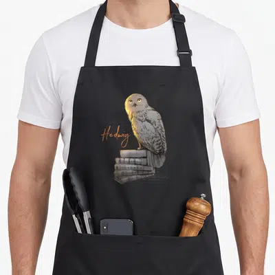 Tablier de cuisine noir avec illustration d'un hibou blanc sur des livres et le mot Hedwig en orange