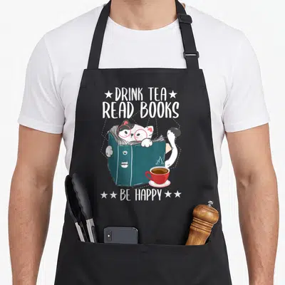 Tablier de cuisine noir avec un chat lisant dans un livre bleu et une tasse de thé rouge.